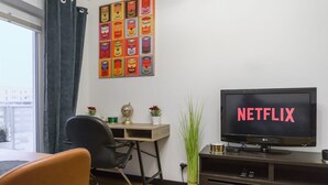 Een woonkamer met een televisie waarop 'NETFLIX' staat, een bureau, een stoel en een muur met een kunstwerk met blikjes.