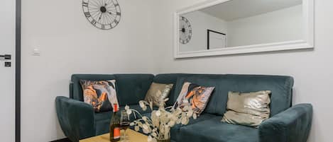 A living room with a dark green sofa, two patterned throw pillows, a small wooden coffee table with a bottle and glasses, and a wall clock.
