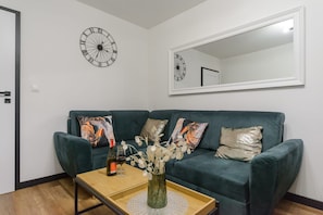 Ein Wohnzimmer mit einem dunkelgrünen Sofa, zwei gemusterten Zierkissen, einem kleinen Holztisch mit einer Flasche und Gläsern sowie einer Wanduhr.