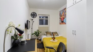 Een kleine slaapkamer met een bed, nachtkastje, lamp en een schilderij aan de muur.