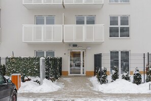 Un immeuble à plusieurs étages avec une entrée centrale, une zone délimitée par une clôture et recouverte de neige, et une voiture garée.