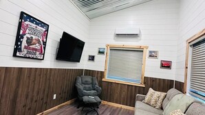 Een kamer met een spartoetziel, een wandgemonteerde flatscreen-tv, een raam met jaloezieën, een grijze bank met gekleurde kussens en een grijze fauteuil.
