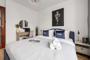 A hotel room with a bed, a desk, a chair, a mirror, a wall art piece, and a bedside table with books and a lamp. - Warsaw Flat near Old Town (Warszawa)