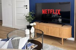 A living room with a television displaying 'NETFLIX' in red letters, a glass coffee table with magazines, a white vase with flowers, and a wooden cabinet. - Pretty House by the Sea for 4 (Łódź)