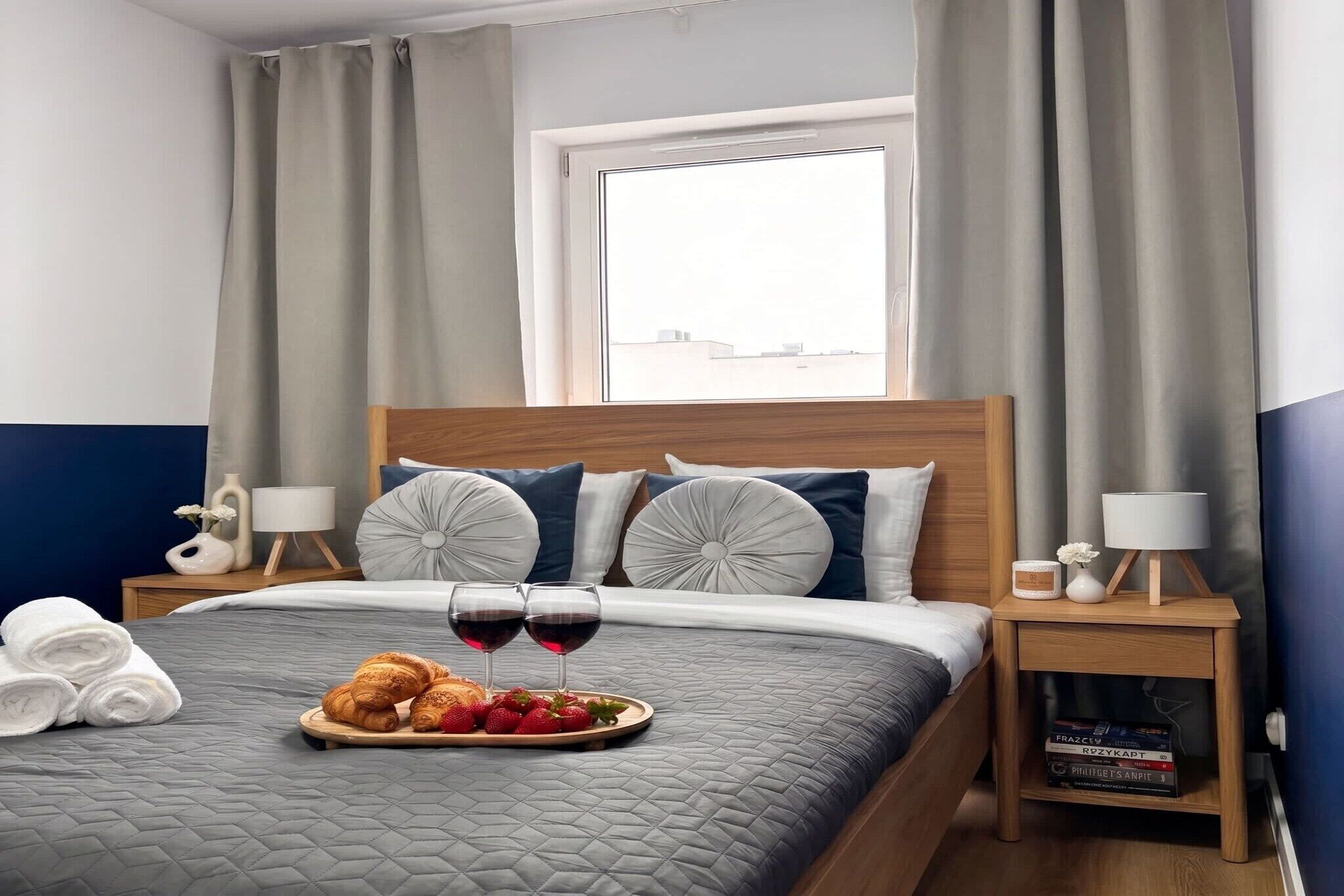 A hotel room with a bed, two bedside tables, a window with curtains, and a tray with croissants and strawberries.