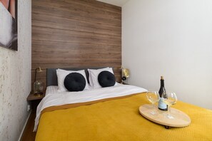 Ein Hotelzimmer mit einem Bett, einem Holz-Kopfstück, zwei Nachttischen mit Lampen, einer gelben Tagesdecke und einem Tablett mit Flasche und Gläsern.