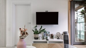 A modern living room with a flat-screen TV mounted on the wall, a white console table with decorative items, a pink vase with flowers, and a large window with a view of buildings. - City Apartment with Balcony (Warszawa)