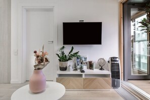 A modern living room with a flat-screen TV mounted on the wall, a white console table with decorative items, a pink vase with flowers, and a large window with a view of buildings. - City Apartment with Balcony (Warszawa)