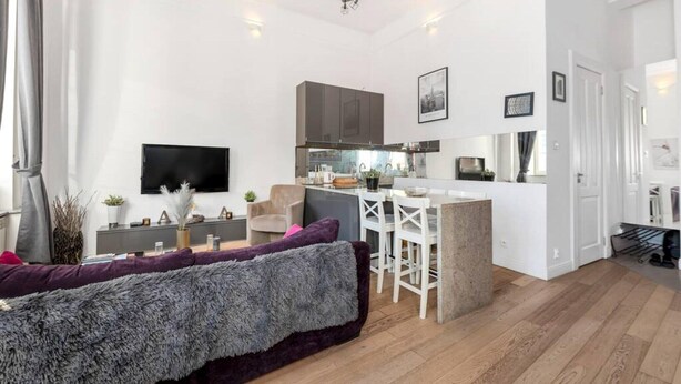 A modern living room with a sofa, a flat-screen TV, and a kitchen area with cabinets and a dining table. - Comfortable house with splendid sea view (Warszawa)