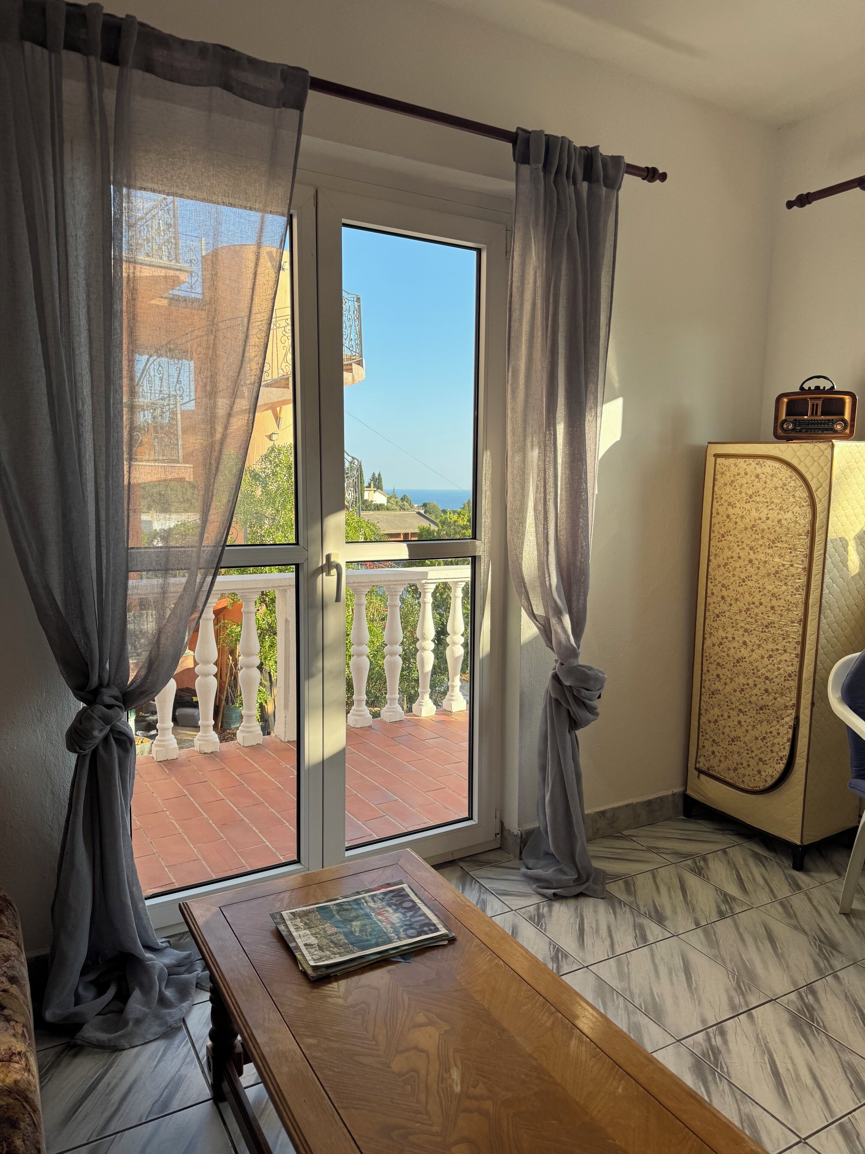 A room with a wooden table, a sofa, a cabinet, and a view of the sea through a glass door.