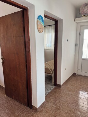 Une pièce avec une porte en bois, un lit avec un cadre blanc, un mur orné d’une décoration et une fenêtre avec des rideaux.
