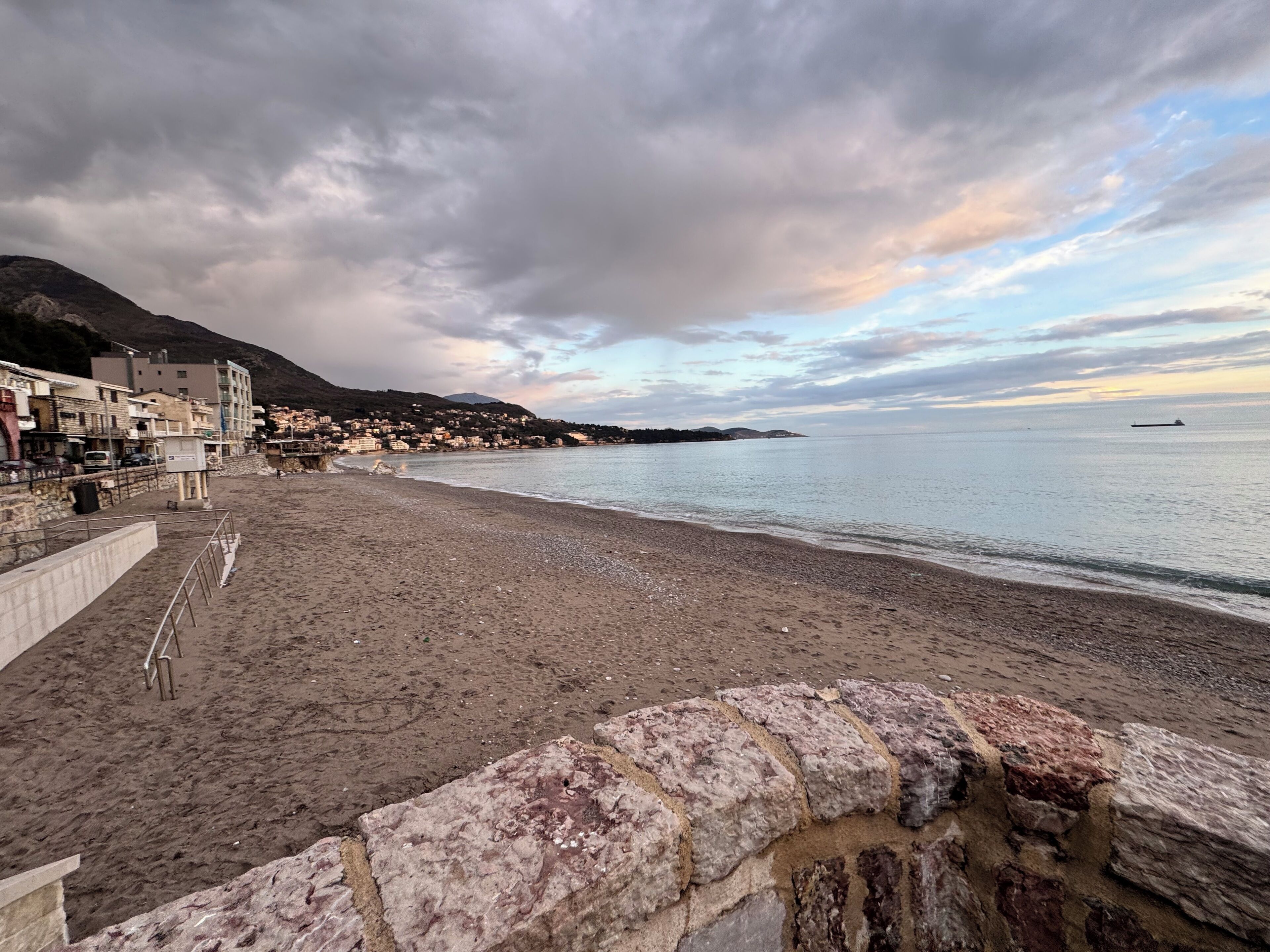 A coastal beach with a stone wall, a clear sky, and a distant view of a town.