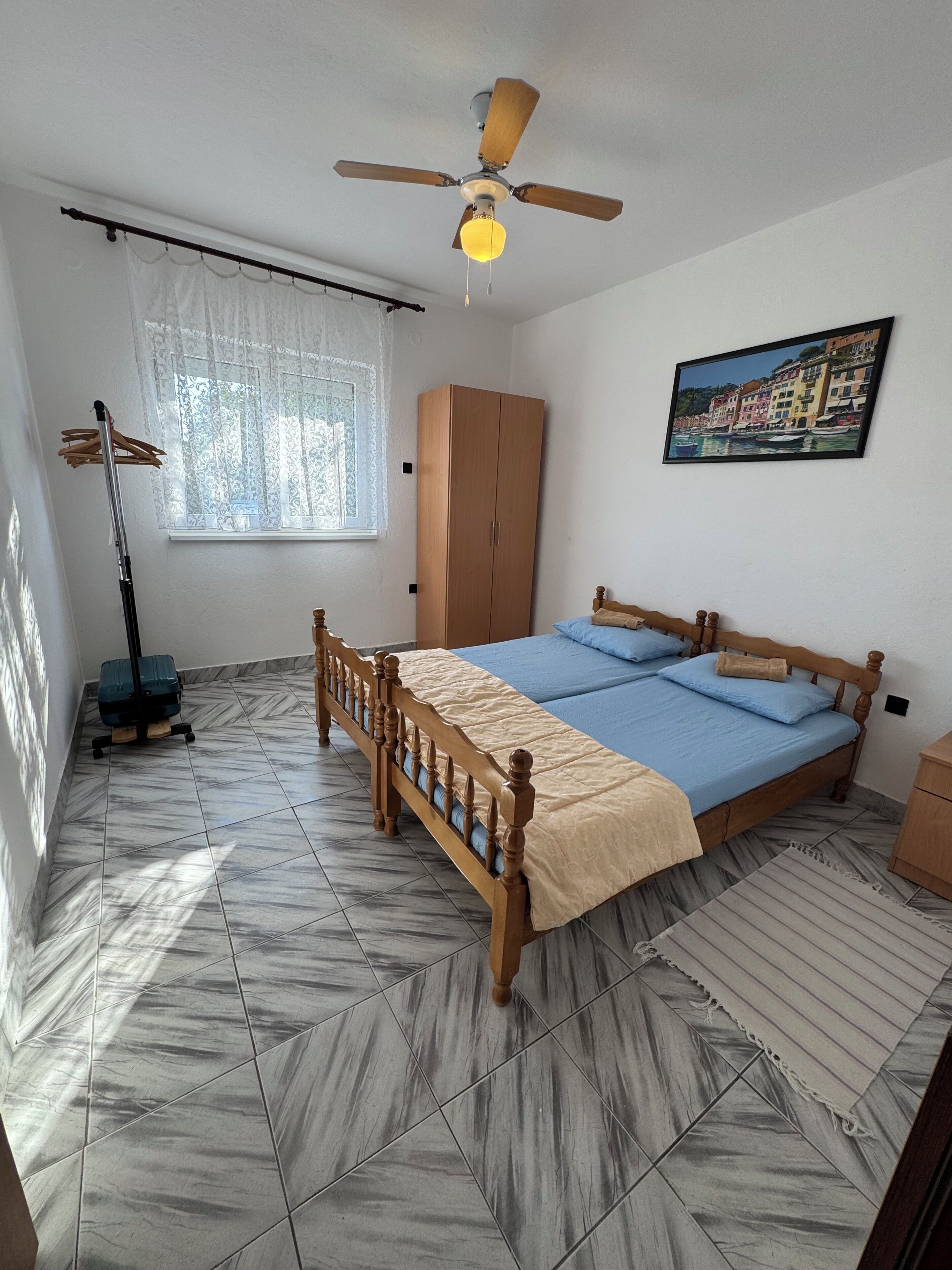 A bedroom with a bed, a wardrobe, a ceiling fan, a painting, and a window with curtains.