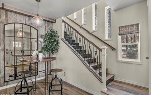 A staircase with a wooden handrail and black balusters, leading to a landing with a round table and stools, a potted plant, and a decorative mirror.