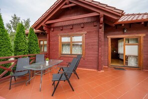 Eine Holzhütte mit einer Terrassenbereiches, ausgestattet mit einem Glastisch und Stühlen, einer Tür zu einem Innenraum und Blick auf den umliegenden Wald.