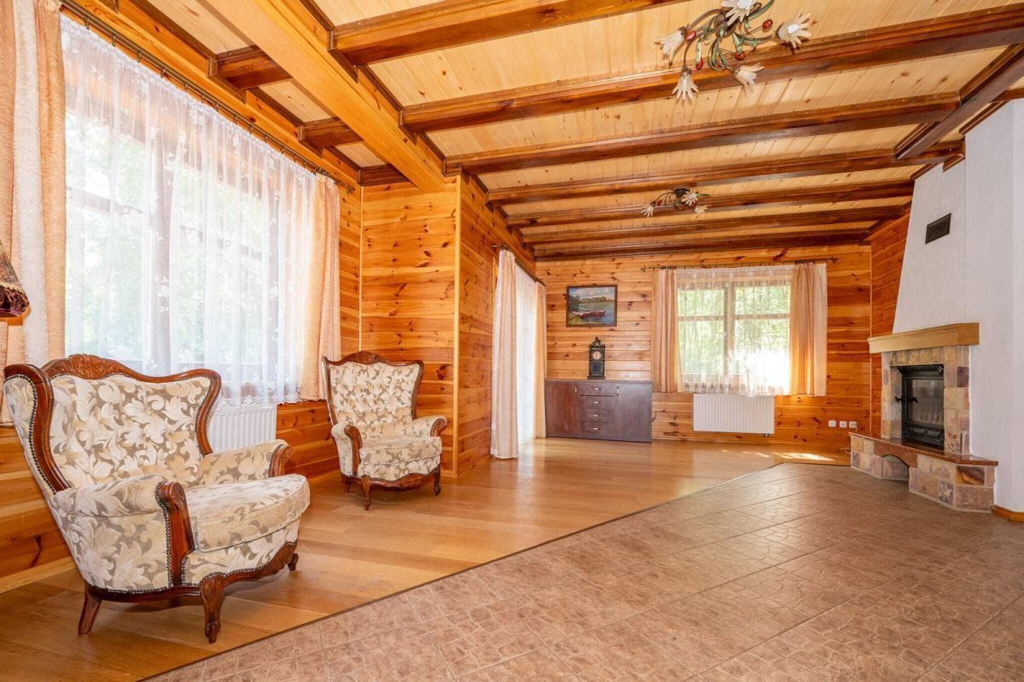 A spacious living room with wooden walls and a high ceiling, featuring two upholstered chairs, a fireplace, and a kitchen area in the background.