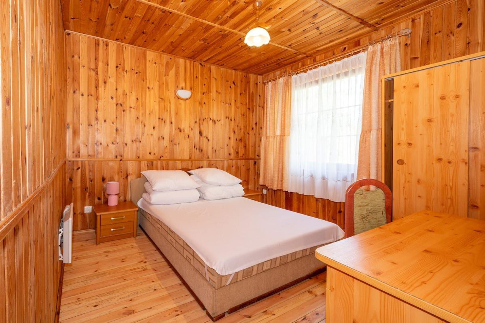 A wooden-paneled room with a bed, a bedside table, a lamp, and a window with curtains.