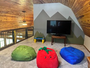 A room with a television on a stand, a wooden ceiling, a green bean bag, a red bean bag, and a blue bean bag. - 5-Acre Private Cabin in the Woods with Kids’ Playroom - No Neighbors in Sight (Mauston)