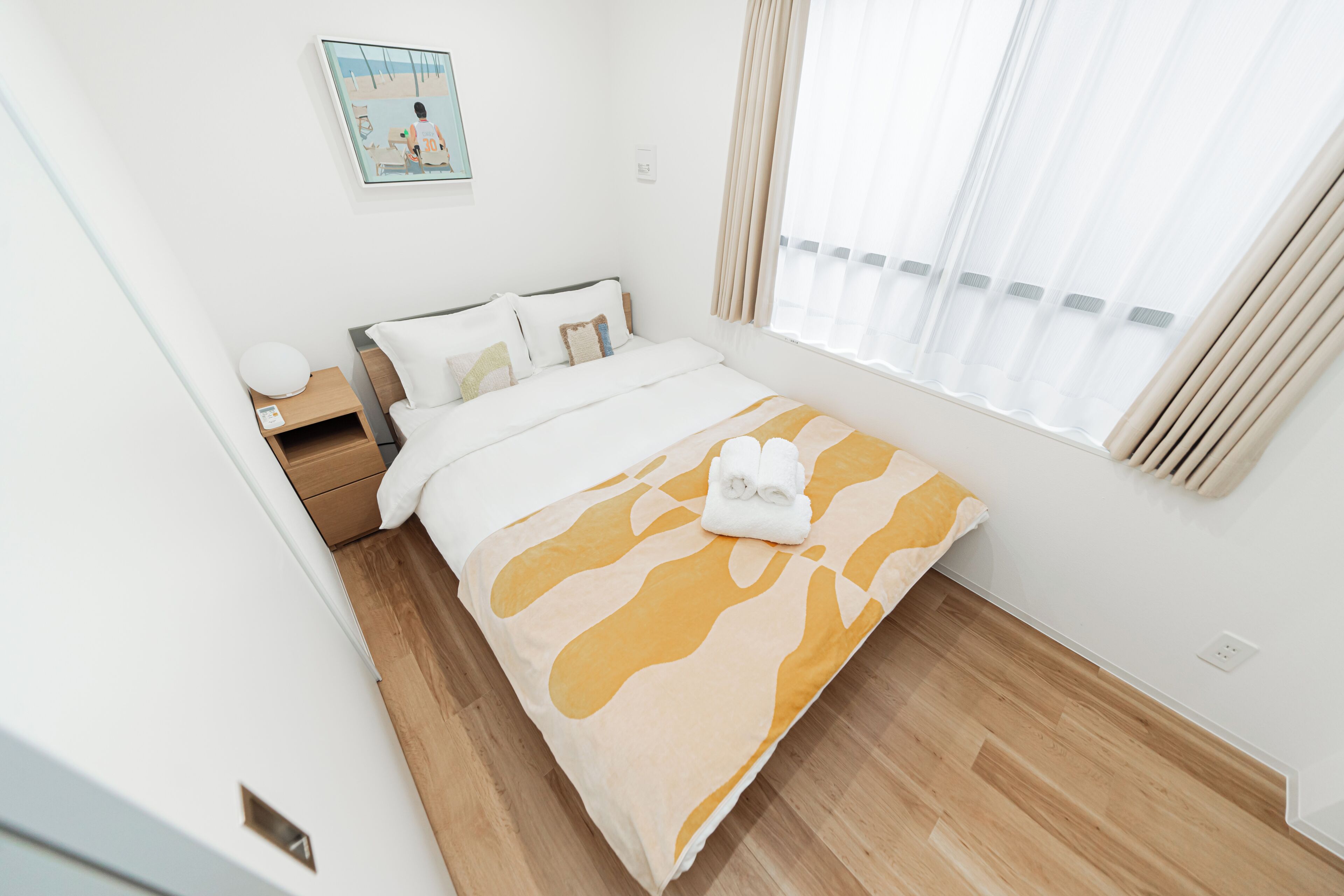 A hotel room with a bed, a nightstand, a lamp, a window with curtains, and a painting on the wall.