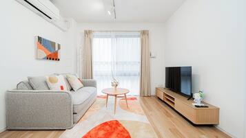 A modern living room with a grey sofa, a round coffee table, a TV on a wooden stand, and a large window with curtains.