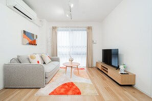 A modern living room with a grey sofa, a round coffee table, a TV on a wooden stand, and a large window with curtains. - Toka Art Hotel (Taito)