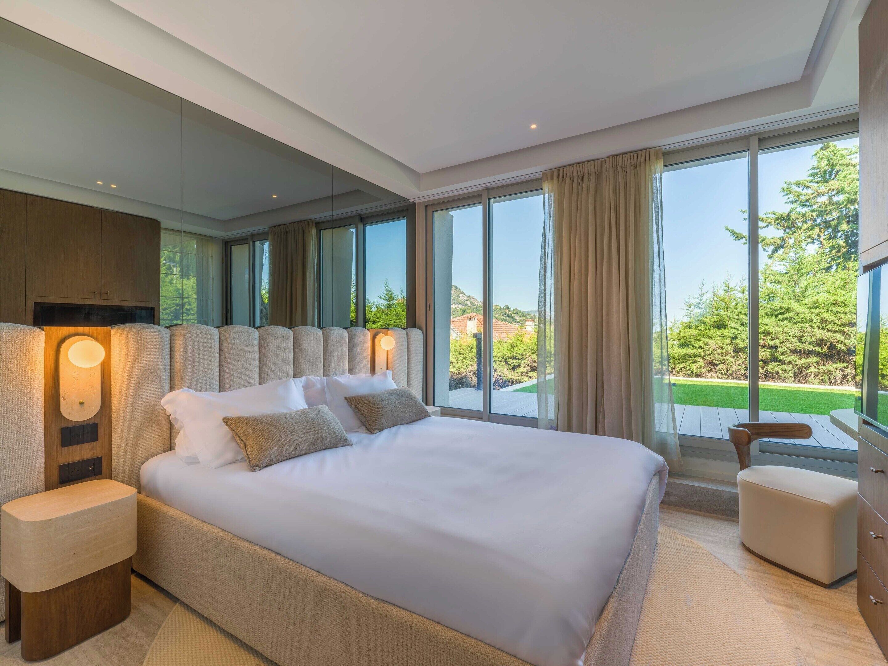 Une chambre moderne avec un grand lit, une table de chevet, une tête de lit en bois et une vue sur un paysage verdoyant grâce à de grandes fenêtres.