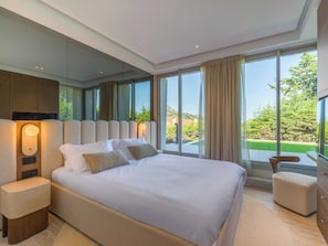 Ein modernes Schlafzimmer mit einem großen Bett, einem Nachttisch, einem Holz-Kopfteil und Blick auf eine grüne Landschaft durch große Fenster.