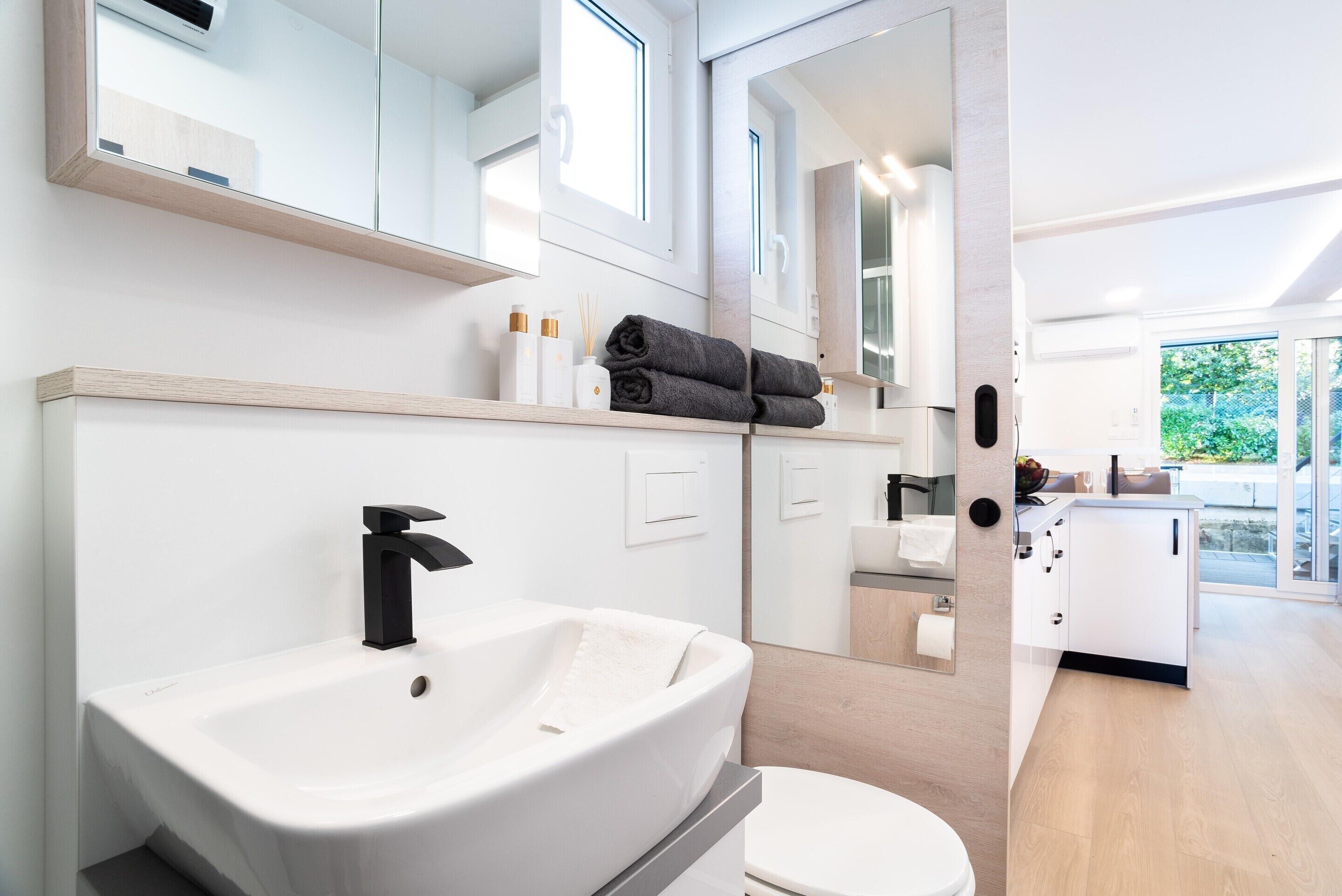A modern bathroom with a large white sink, a black faucet, and a toilet. There are towels and toiletries on the countertop.