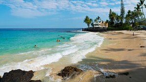 A beach with clear turquoise water, a rocky shoreline, and a building with a balcony overlooking the ocean. - Newly Renovated Oceanfront Condo at Magic Sands Beach (Kailua-Kona)