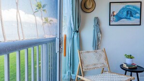 A balcony with a view of palm trees, a woven chair, a small table with a plant, and a framed picture on the wall. - Newly Renovated Oceanfront Condo at Magic Sands Beach (Kailua-Kona)