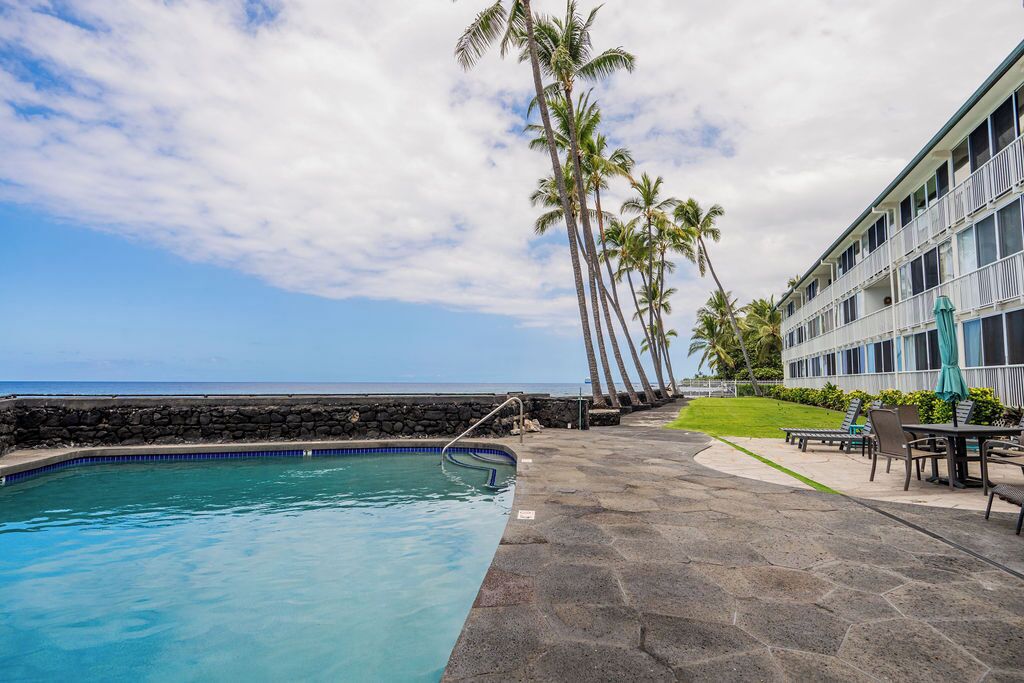 A pool area with a stone deck, lounge chairs, and a building with balconies.
