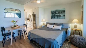 A bedroom with a bed, bedside tables, a desk, chairs, a mirror, and a view of the kitchen. - Newly Renovated Oceanfront Condo at Magic Sands Beach (Kailua-Kona)