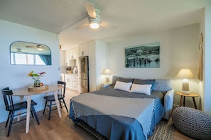 Une chambre à coucher comprenant un lit, des tables de chevet, un bureau, des chaises, un miroir et donnant sur la cuisine.