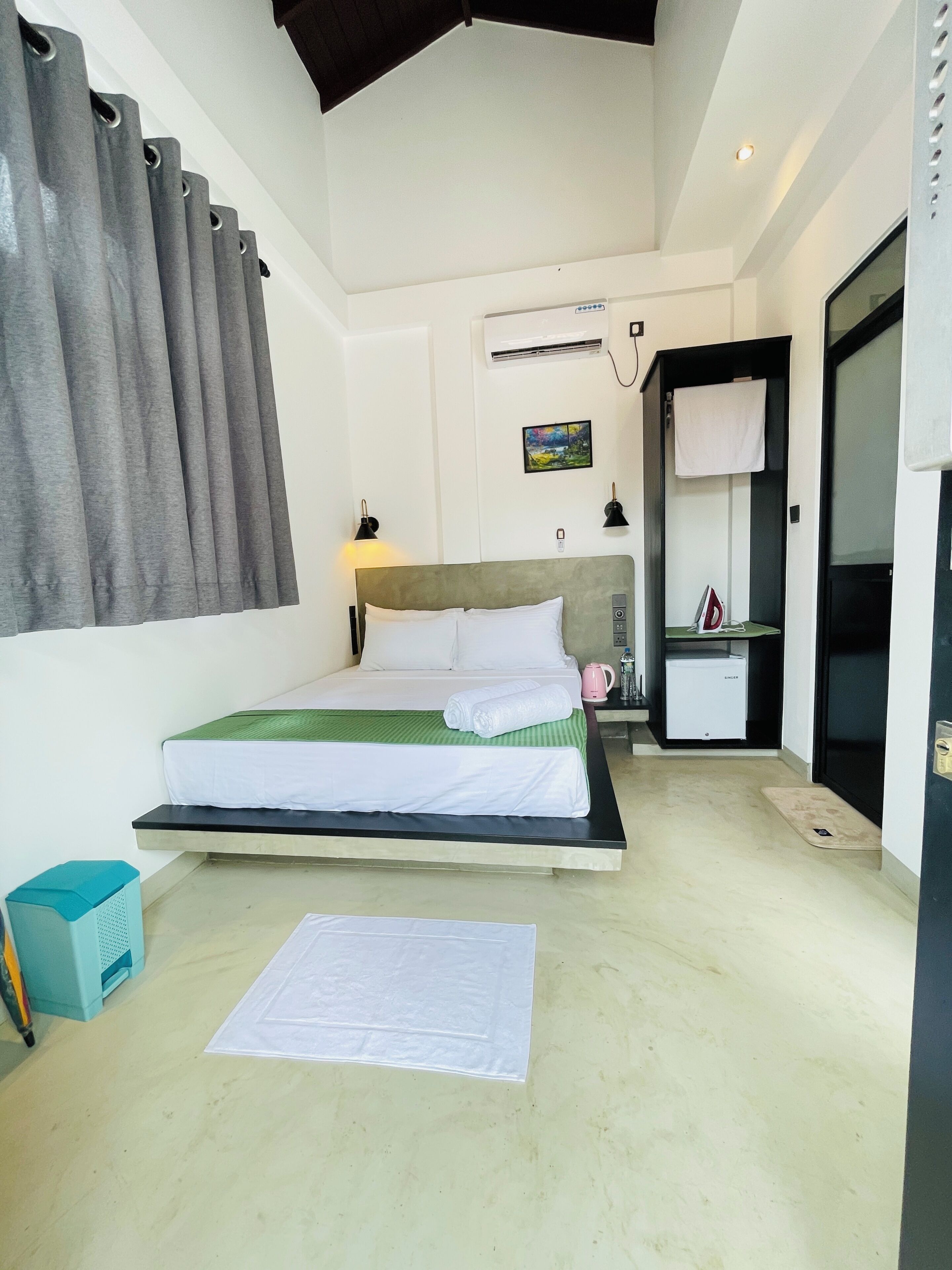 Une chambre d’hôtel comprenant un lit, un bureau, une chaise, une petite table, un radiateur, une bouilloire, un tableau au mur et une fenêtre avec des rideaux.