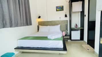 Une chambre d’hôtel comprenant un lit, un bureau, une chaise, une petite table, un radiateur, une bouilloire, un tableau au mur et une fenêtre avec des rideaux.