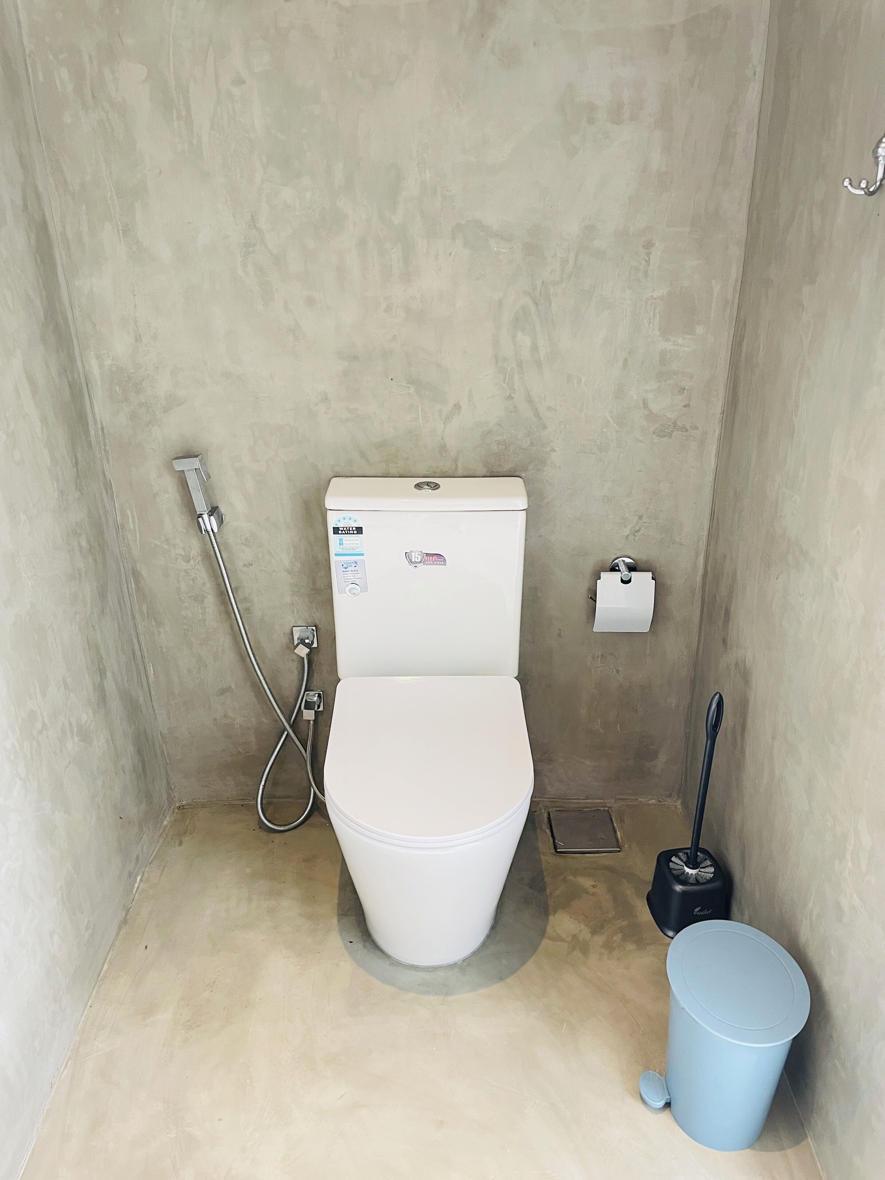 A small, compact bathroom with a white toilet, a handheld showerhead, a toilet brush, and a trash bin.