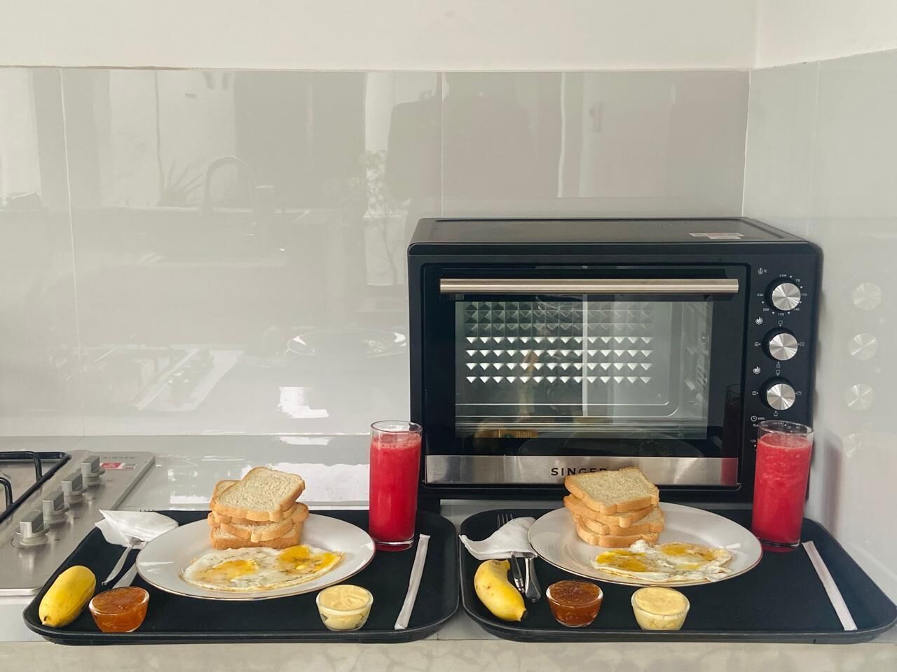 Una cocina con horno tostador, dos platos con tostadas y huevos, un plátano y dos vasos de jugo rojo.