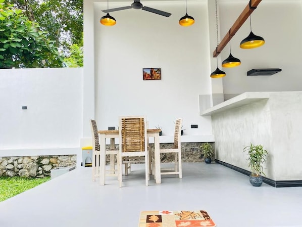 Un área de comedor al aire libre con una mesa, sillas, un ventilador de techo, luces colgantes y una planta en maceta.