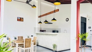 Comedor con una mesa y sillas blancas, ventilador de techo, luces colgantes y una pequeña planta en maceta.