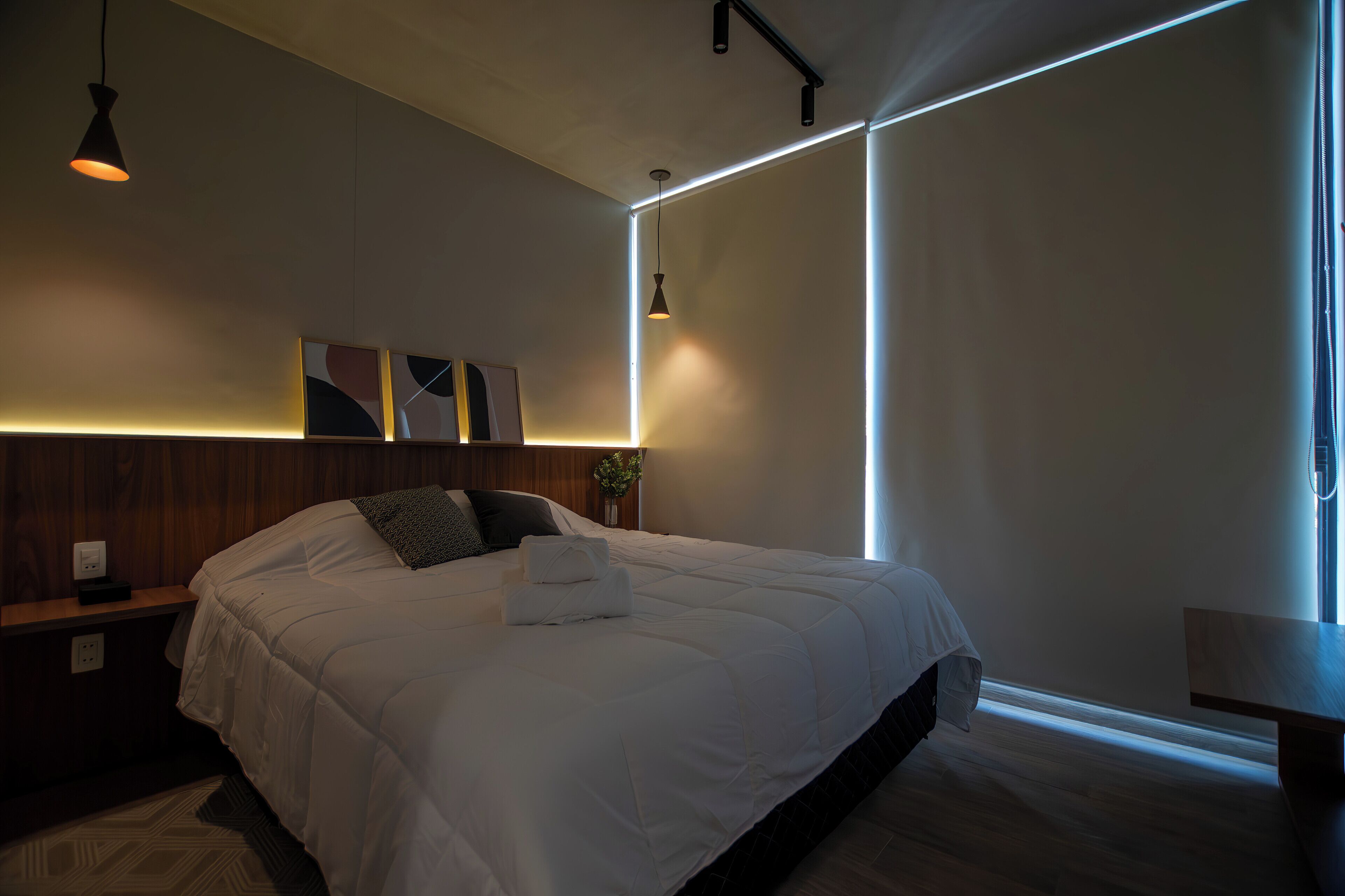 Une chambre moderne avec un grand lit, des tables de chevet, une petite table d’appoint, une lampe fixée au mur et une fenêtre avec des rideaux.