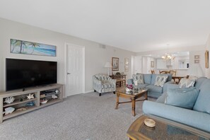 A living room with a flat-screen TV, a sofa, a coffee table, and a dining area with a table and chairs. - ☀️Beach Front-Sunset Views | Pool and Easy Beach Access | Sand Caper #702 🌴 (Fort Myers Beach)