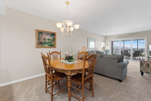 A dining area with a round table, chairs, a chandelier, and a painting on the wall. - ☀️Beach Front-Sunset Views | Pool and Easy Beach Access | Sand Caper #702 🌴 (Fort Myers Beach)