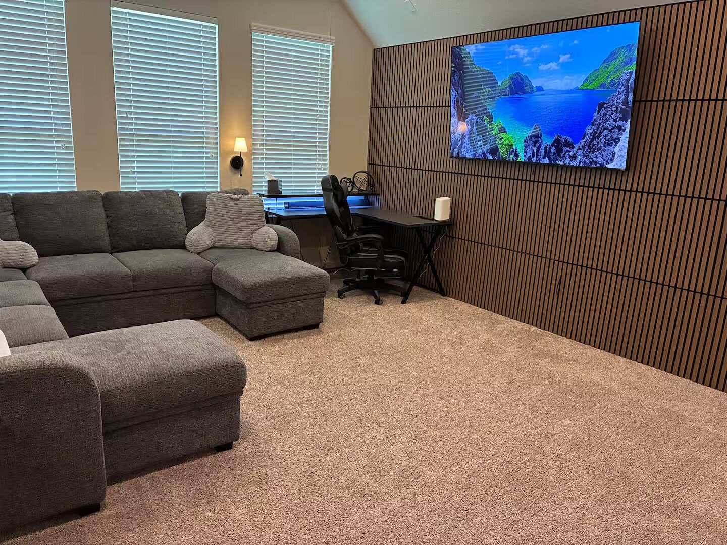 Ein Wohnzimmer mit einem Ecksofa, einem Schreibtisch mit Stuhl und einem großen Wandfernseher.