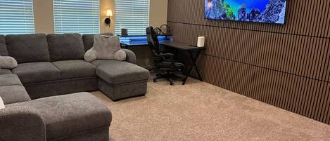 Ein Wohnzimmer mit einem Ecksofa, einem Schreibtisch mit Stuhl und einem großen Wandfernseher.