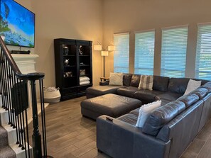 A living room with a sectional sofa, a black entertainment unit, a lamp, and a TV on the wall. - Your Sleep Destination Awaits (Cypress)