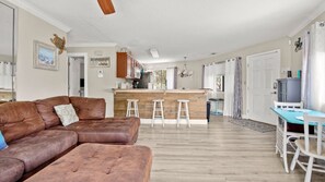 Ein Wohnzimmer mit braunem Ledersofa, einer Küchenzeile mit Holzausstattung und einem Esstisch mit weißen Stühlen.