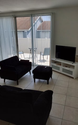 A living room with a sofa, ottoman, TV stand, and a sliding glass door leading to a balcony. - Perfect Getaway Near Disney & Universal (Davenport)