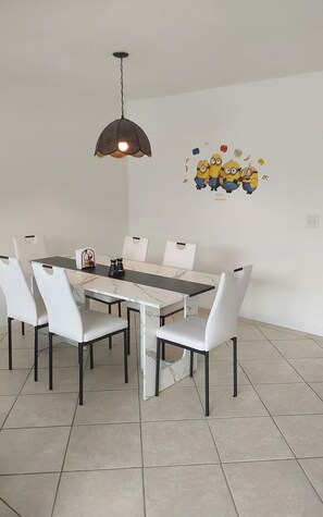 Un coin repas avec une table surmontée d’un plateau en marbre, des chaises blanches, un luminaire suspendu et un autocollant mural représentant des personnages de dessins animés.