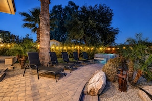 Un área de estar al aire libre con sillones reclinables y un jacuzzi, rodeada de árboles y luces decorativas.