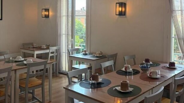 A dining area with multiple tables and chairs, a hanging light fixture, and a view through the window. - Hôtel du Commerce (Millau)